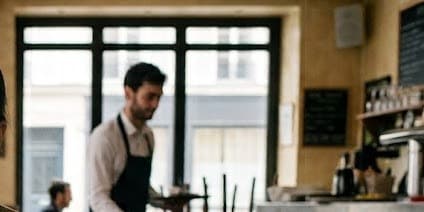 Un serveur en tablier derrière le comptoir d'une brasserie, devant la porte vitrée qui donne sur la rue.