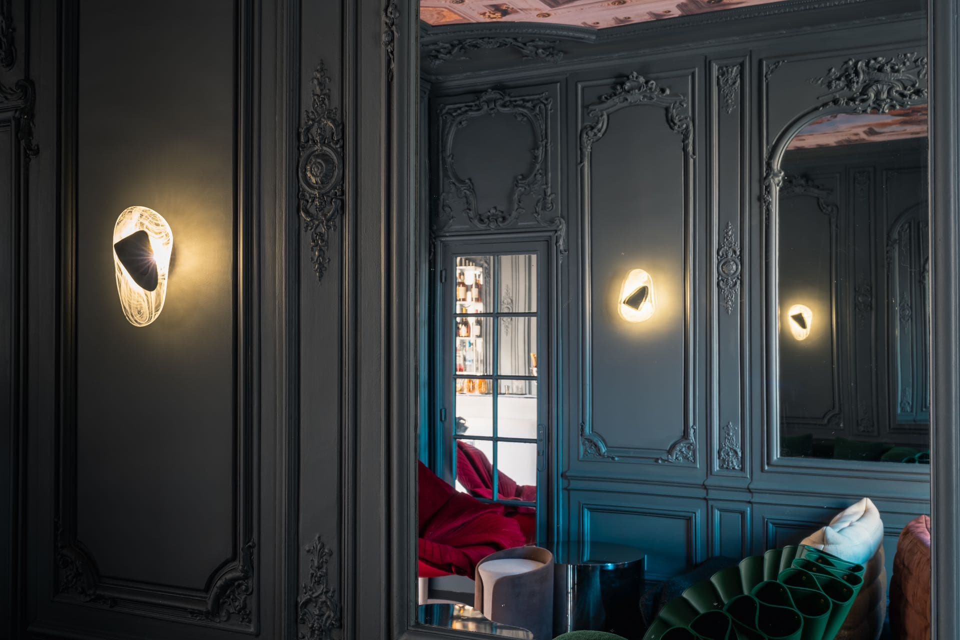 Appliques lumineuses sur boiseries grises ornées, miroir et fauteuils rouge et vert dans un salon sombre